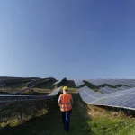 Técnico caminhando entre fileiras de painéis solares em uma usina fotovoltaica, cenário típico de inspeções e diagnósticos realizados durante serviços de consultoria em energia solar em projetos de grande porte.