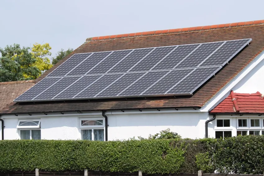 Módulo fotovoltaico instalado em telhado marrom de casa com paredes brancas, rodeada por uma cerca viva