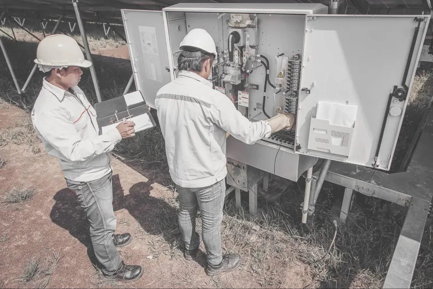 Dois homens em pé, vestindo roupas casuais e equipamentos de proteção, em frente a uma string box aberta. Um deles está com ambas as mãos na string box, enquanto o outro faz anotações em uma planilha de papel