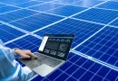 Pessoa utilizando notebook com sistema de monitoramento de energia solar em tempo real, em frente a painéis solares azuis.